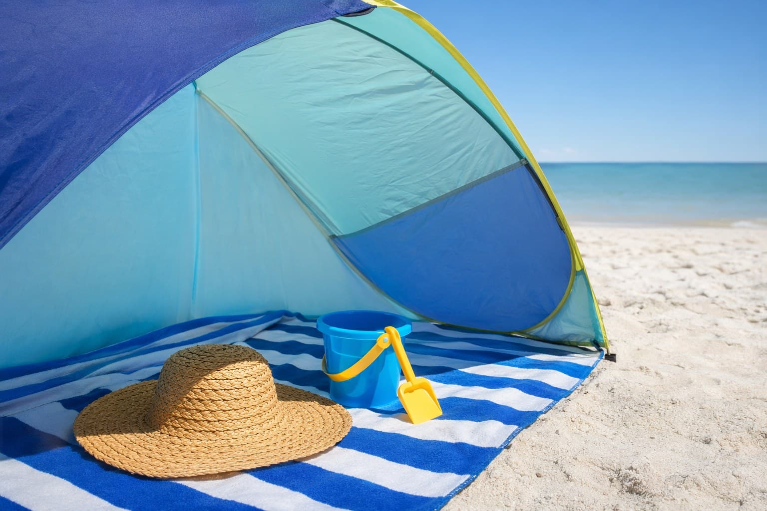 A striped pop-up beach canopy casting cool shade over a blanket and a sun hat on pale sand