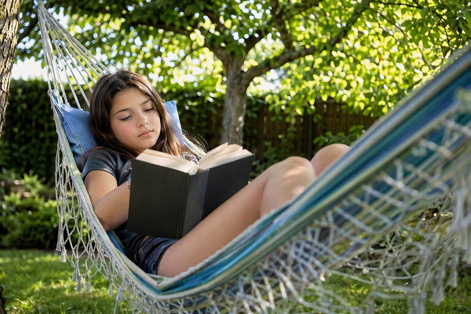 A tween reading independently in a hammock outdoors on a sunny afternoon, fully absorbed in a thick novel